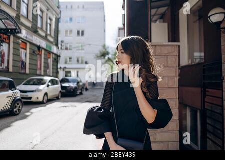 Bella ragazza modello in abito da sera, in piedi per la strada e raddrizzando i capelli Foto Stock