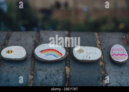 Londra, UK - 12 aprile 2020: Ciottoli dipinti con arcobaleno, uovo di Pasqua, stare al sicuro e grazie messaggi NHS lasciato sulla strada come la gente sostiene ciascuno Foto Stock