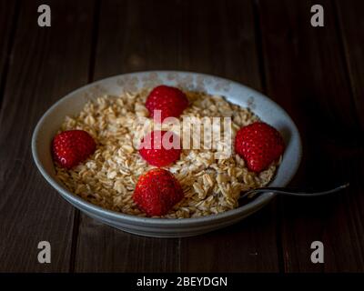 Farinata d'avena in un piatto marrone decorato con fragole dimezzate su tavolo di legno, piatto intero, centrato, vista dall'alto, con cucchiaio di argenteria Foto Stock