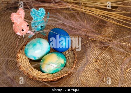 Uova pasquali multicolore dai toni blu e due coniglietti pasquali su un bastone su uno sfondo di stoffa marrone. Foto Stock