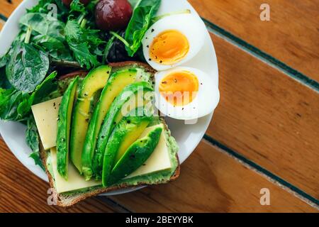 Toast con avocado, uova bollite, spinaci e pomodori su piatto bianco con coltello e forchetta su serviette e rustico sfondo in legno.