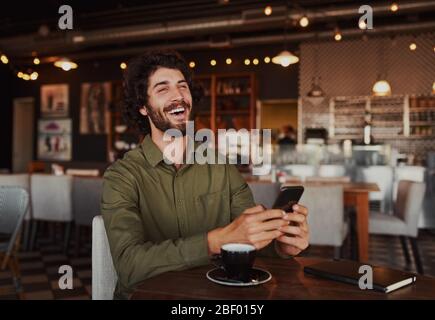 Ritratto di bel giovane uomo ridente mentre legge il messaggio divertente al telefono seduto in caffetteria durante il tempo libero Foto Stock