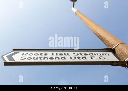Cartello direzionale a livello della strada che punta a sinistra verso il Roots Hall Stadium, sede del Southend United Football Club, sotto il cielo azzurro. Foto Stock