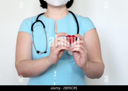 Salvare vite durante una pandemia di coronavirus 19 covid, medico della donna in maschera medica che tiene il cuore rosso lavorato a maglia nelle mani. Concetto di cardiologo Foto Stock