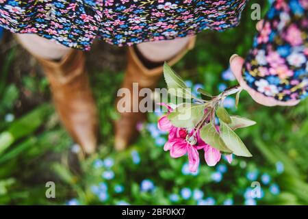 Una donna o una ragazza che si trova all'esterno in una giornata primaverile indossando un abito floreale e stivali cowboy e tenendo rosa fioritura, fresco e floreale. Dof poco profondo. Foto Stock
