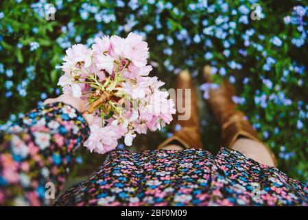 Una donna o una ragazza che si trova all'esterno in una giornata primaverile indossando un abito floreale e stivali cowboy e tenendo rosa fioritura, fresco e floreale. Dof poco profondo. Foto Stock