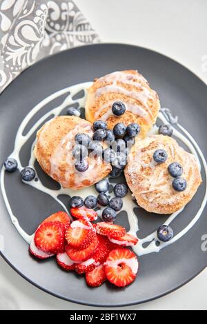 Pancake o sciroppi di formaggio casolare con mirtillo e fragole su piatto nero, vista primo piano. Salsa di latte condensato. Cucina russa, Ucraina Foto Stock