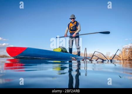 Senior maschio pagatore su una stand up paddleboard, un lago tranquillo in Colorado, inverno o primi paesaggi primaverili, vista bassa angolazione fotocamera, ricreazione, fitness Foto Stock