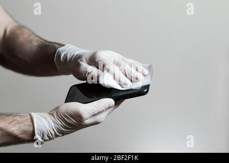 A mans mani che indossano guanti in lattice strofinando il suo cellulare con una salvietta disinfettante durante la pandemia Covid 19. Foto Stock