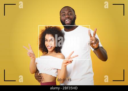 Buon blogger afro-americano che mostra il gesto della vittoria mentre registra video su sfondo a colori Foto Stock