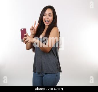 Una giovane donna carina in jeans e una camicia grigia tiene il segno di pace come lei prende una foto sul suo telefono cellulare. Foto Stock