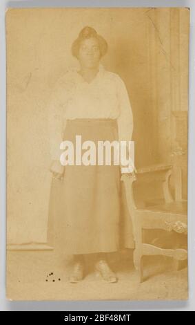 Cartolina fotografica di una donna. Cartolina fotografica di una donna seduta sul braccio di una panca. La donna è vestita con un abito e una gonna in tessuto lucido. La gonna ha un design applicato. È seduta di fronte alla fotocamera, con il braccio sinistro appoggiato sul retro del banco, che è girato lontano dalla fotocamera. Foto Stock