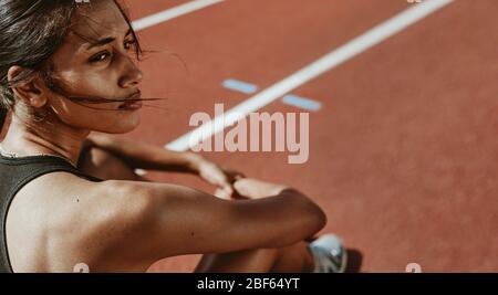 Primo piano di una donna in forma dopo corsa seduta su pista. Atleta determinata che si siede sul campo e guarda lontano. Foto Stock