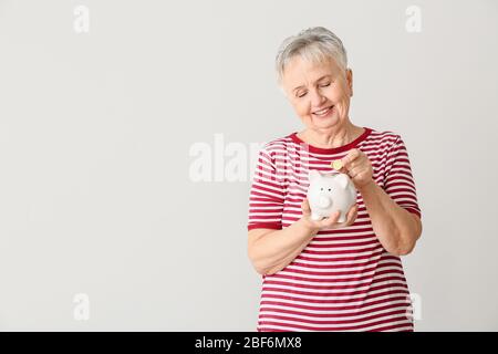 Donna anziana che mette i soldi in banca del piggy su sfondo chiaro Foto Stock