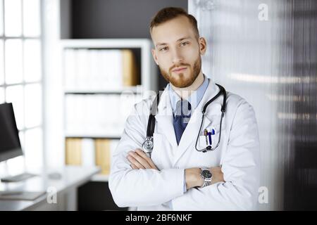 Medico rosso-bearded che sta diritto in clinica vicino al suo posto di lavoro. Ritratto del medico. Medicina e concetto di salute Foto Stock