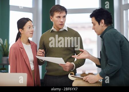 Gruppo di colleghi in piedi e parlando tra loro e discutendo documenti durante il giorno lavorativo in ufficio Foto Stock