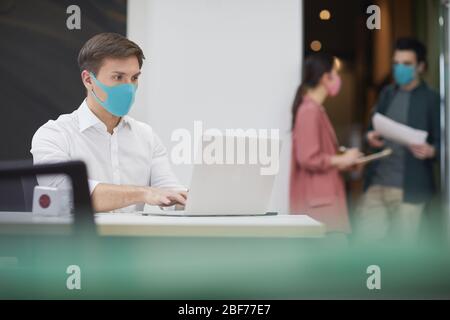 Giovane uomo d'affari in maschera protettiva seduto al tavolo e digitando sul computer portatile in ufficio Foto Stock