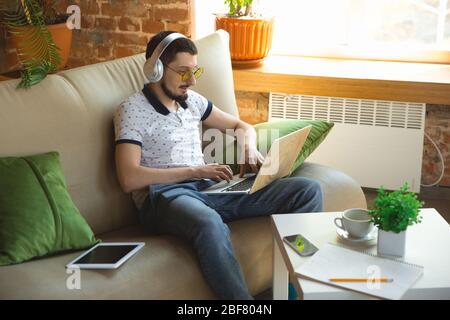 Utilizzo di gadget. Uomo che lavora da casa durante il coronavirus o la quarantena COVID-19, concetto di ufficio remoto. Giovane uomo d'affari, manager che svolge attività con smartphone, computer, ha conferenze online, riunione. Foto Stock