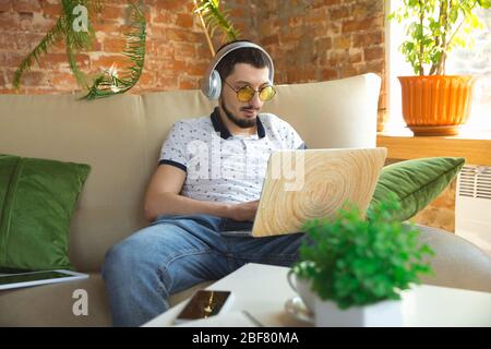 Utilizzo di gadget. Uomo che lavora da casa durante il coronavirus o la quarantena COVID-19, concetto di ufficio remoto. Giovane uomo d'affari, manager che svolge attività con smartphone, computer, ha conferenze online, riunione. Foto Stock