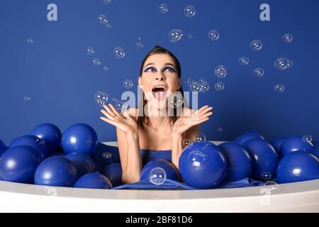 Ritratto di giovane donna alla moda con palloncini d'aria e bolle di sapone seduti nella vasca da bagno su sfondo blu Foto Stock