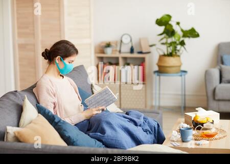 Donna malata che indossa maschera protettiva seduta sul divano con libro è in congedi per malattia e seduta a casa Foto Stock