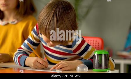 Apprendimento a distanza per gli scolari durante il periodo di quarantena del coronavirus. I bambini sono seduti al tavolo. In primo piano il ragazzo prende appunti a matita. Foto Stock