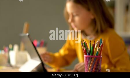 Apprendimento a distanza a casa durante il periodo di quarantena del coronavirus Covid 19. Una ragazza siede a un tavolo e guarda in un notebook. In primo piano è un vetro con matite colorate. Foto Stock