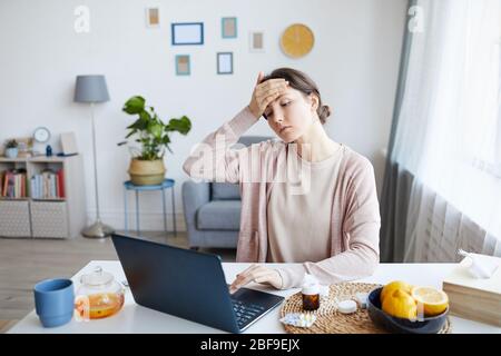 Giovane donna ha una temperatura elevata mentre si siede al tavolo e lavora a casa sul computer portatile Foto Stock