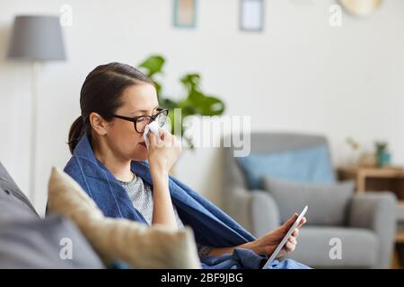 Giovane donna malata negli occhiali ha preso un freddo che riposava sul divano e guardando il film su tablet digitale nel soggiorno a casa Foto Stock