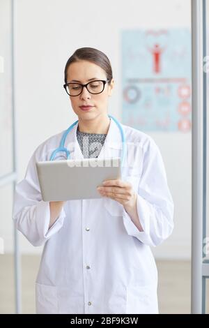 Medico donna serio in occhiali che indossano cappotto bianco e che lavora online su tablet digitale mentre si trova in ufficio Foto Stock