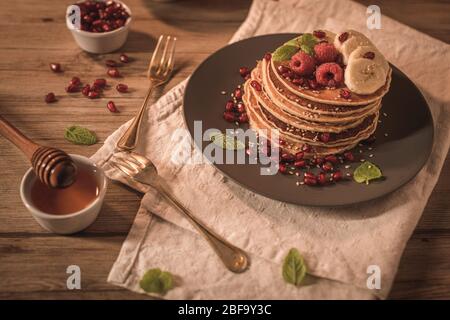 Pancake con lamponi, fette di banana, semi di melograno e miele su tavola d'annata in legno. Foto Stock