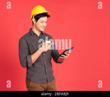 Giovane uomo asiatico bello in abito casual con cappello giallo mano tenendo laptop in architetto e ingegnere lavoratore isolato in sfondo rosso in studio Foto Stock