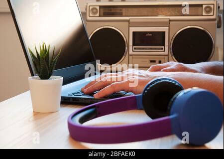 Vista parziale dell'uomo che lavora con il laptop vicino alle cuffie al tavolo. Uomo che digita sul laptop. Le cuffie wireless e un registratore stereo 80s sono sul woode Foto Stock