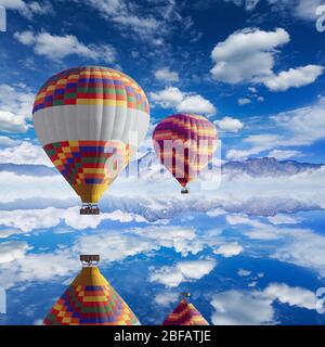 Palloncini colorati ad aria calda vola sopra il lago calmo, nuvole bianche nel cielo blu, alte montagne all'orizzonte Foto Stock