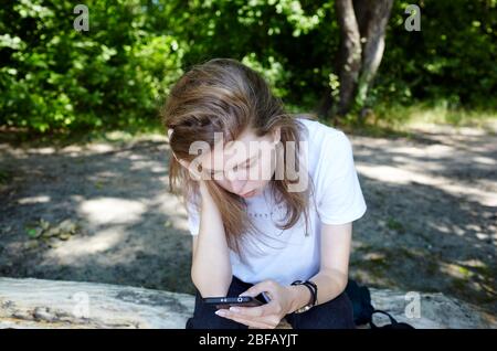 Bella donna attraente in bianco t-shirt in posa nel parco estivo. Ragazza attiva che tiene il telefono cellulare e riposa all'aperto Foto Stock