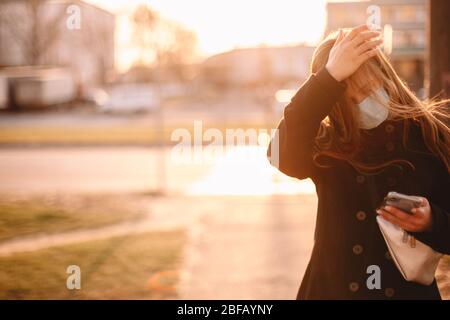 Giovane donna in maschera protettiva viso medico toccare la testa mentre si cammina in strada in città utilizzando lo smartphone Foto Stock