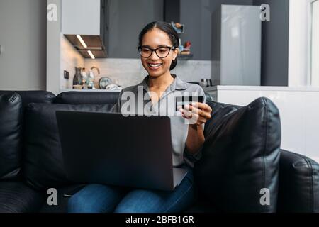 Donna sorridente che acquista online con carta di credito a casa sul divano utilizzando il laptop Foto Stock