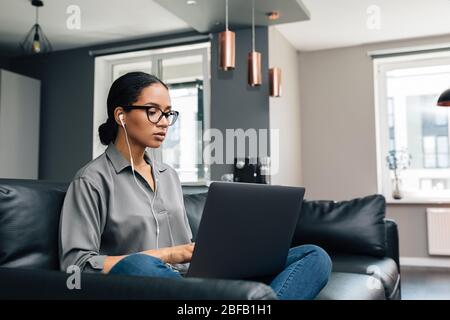 Giovane donna che lavora dal suo appartamento. Freelancer femminile che usa un laptop seduto sul divano. Foto Stock