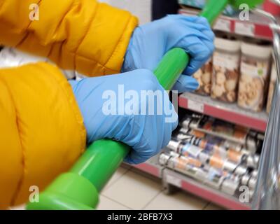 Le mani donna in guanti protettivi per uso medico tengono in primo piano l'impugnatura del carrello della spesa. Protezione del coronavirus nel supermercato Foto Stock