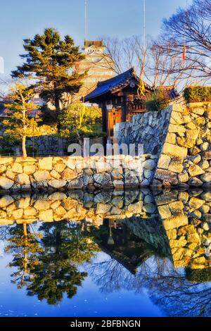 Muri di pietra intorno al parco storico nella città di Himeji del Giappone all'alba con alta torre bianca che riflette nelle acque del fossato. Foto Stock