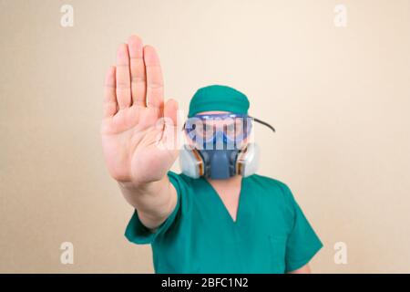 Medico maschile in tuta verde e maschera respiratore con occhiali protettivi fa SEGNO DI STOP con la sua mano. Messa a fuoco selettiva. Concetto medico. Foto Stock