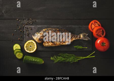 Pesce di orata con pomodori, cetrioli, aneto e una fetta di limone su una pietra nera su sfondo nero ligneo Foto Stock