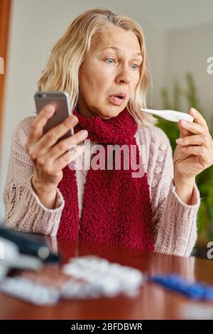 La donna anziana, che ha problemi, tiene un termometro nelle mani e chiama un telefono Foto Stock