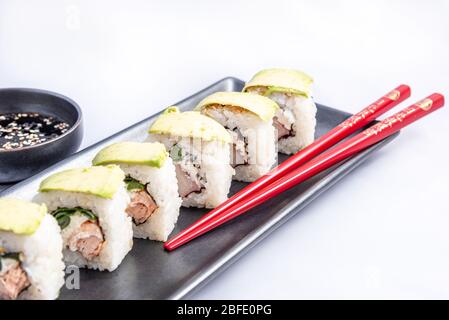 Rotolo di sushi con wrap avocado, salmone alla griglia, formaggio philadelphia e cipolle verdi contenitore nero e bacchette rosse. Foto Stock