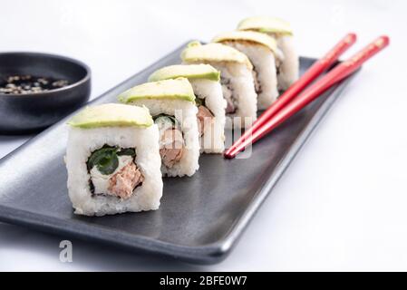 Rotolo di sushi con wrap avocado, salmone alla griglia, formaggio philadelphia e cipolle verdi contenitore nero e bacchette rosse. Foto Stock