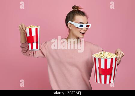 giovane bella donna caucasica che indossa occhiali rosso-blu 3d e tenendo due secchi di popcorn isolato su sfondo rosa. ragazza sceglie tra grande e piccolo secchio di popcorn Foto Stock