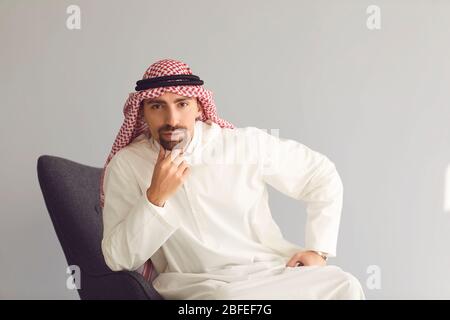 L'uomo arabo serio sembra seduta mentre si siede su una poltrona su sfondo grigio Foto Stock