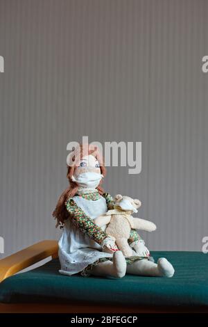 Una bambola femminile fatta a mano e in vecchio stile vestita con un abito con un pinafora è seduta su una sedia, indossando una maschera protettiva. Lei tiene un piccolo orso di teddy Foto Stock