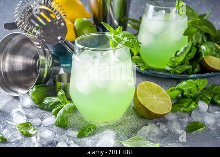 Cocktail alcolico con gin di basilico. Ricetta lunga bevanda alcolica con foglie di basilico fresco e lime, spazio copia sfondo pietra grigia Foto Stock