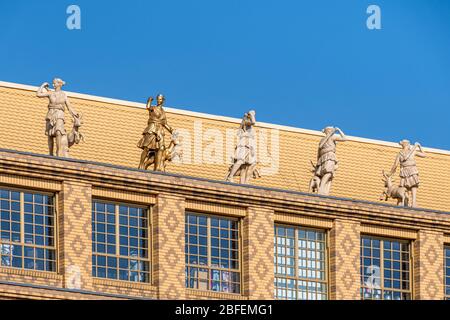 Statua dorata oltre a statue di marmo sulla parte superiore della facciata di mattoni gialli con motivi e grandi finestre Foto Stock